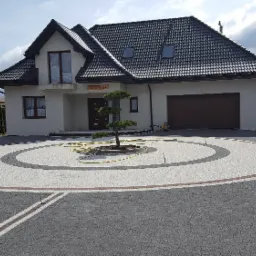 Nowoczesny dom z podjazdem wyłożonym kostką w odcieniach szarości i bieli, z okrągłym wzorem i małym drzewkiem pośrodku, widoczny fragment żółtego węża ogrodowego.