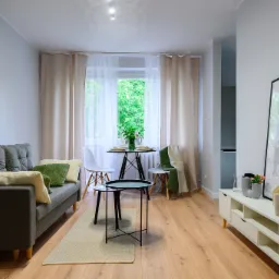 Jasne wnętrze salonu po remoncie: szara sofa z poduszkami, drewniana podłoga, biała komoda z dekoracjami, okno z zasłonami i okrągły stół z krzesłami.