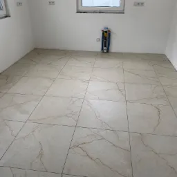 Jasne, kwadratowe płytki podłogowe z marmurkowym wzorem w nowym pomieszczeniu, widoczne instalacje hydrauliczne przy ścianie i butelka wody w rogu.