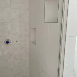 Niedokończona łazienka z białymi płytkami z delikatnym wzorem, wnękami w ścianie i zaklejonym otworem na baterię.