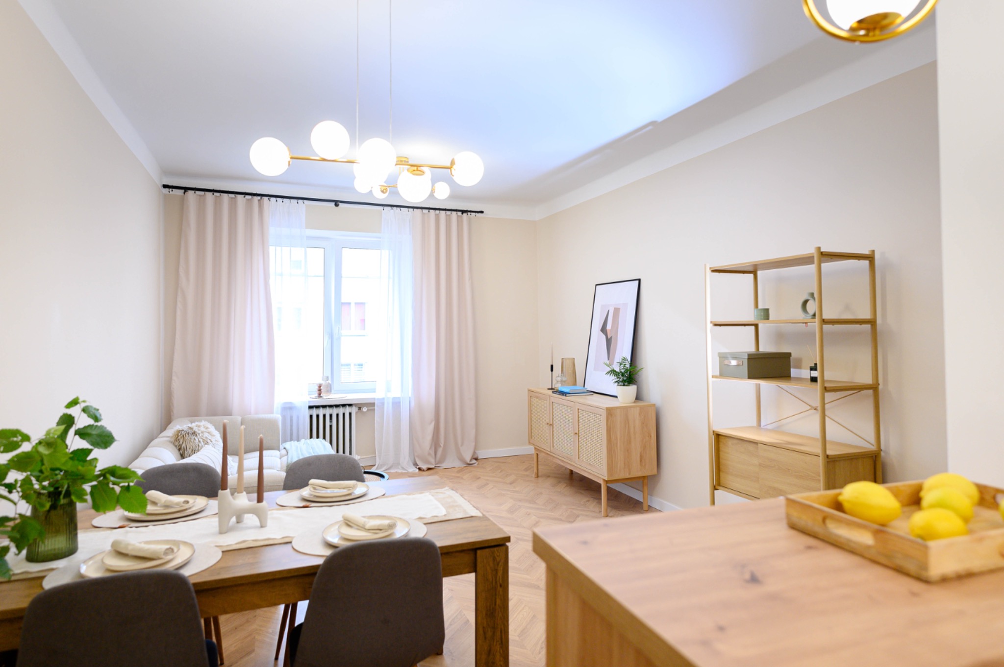 Jasne wnętrze jadalni z drewnianym stołem, krzesłami w odcieniach szarości i stylową komodą. Na podłodze parkiet ułożony w jodełkę. Oświetlenie sufitowe w stylu retro.