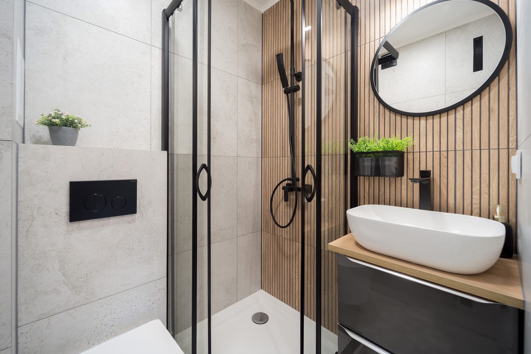 Nowoczesna łazienka z kabiną prysznicową typu walk-in, umywalką nablatową i drewnianymi panelami ściennymi. Czarna armatura i akcenty roślinne.