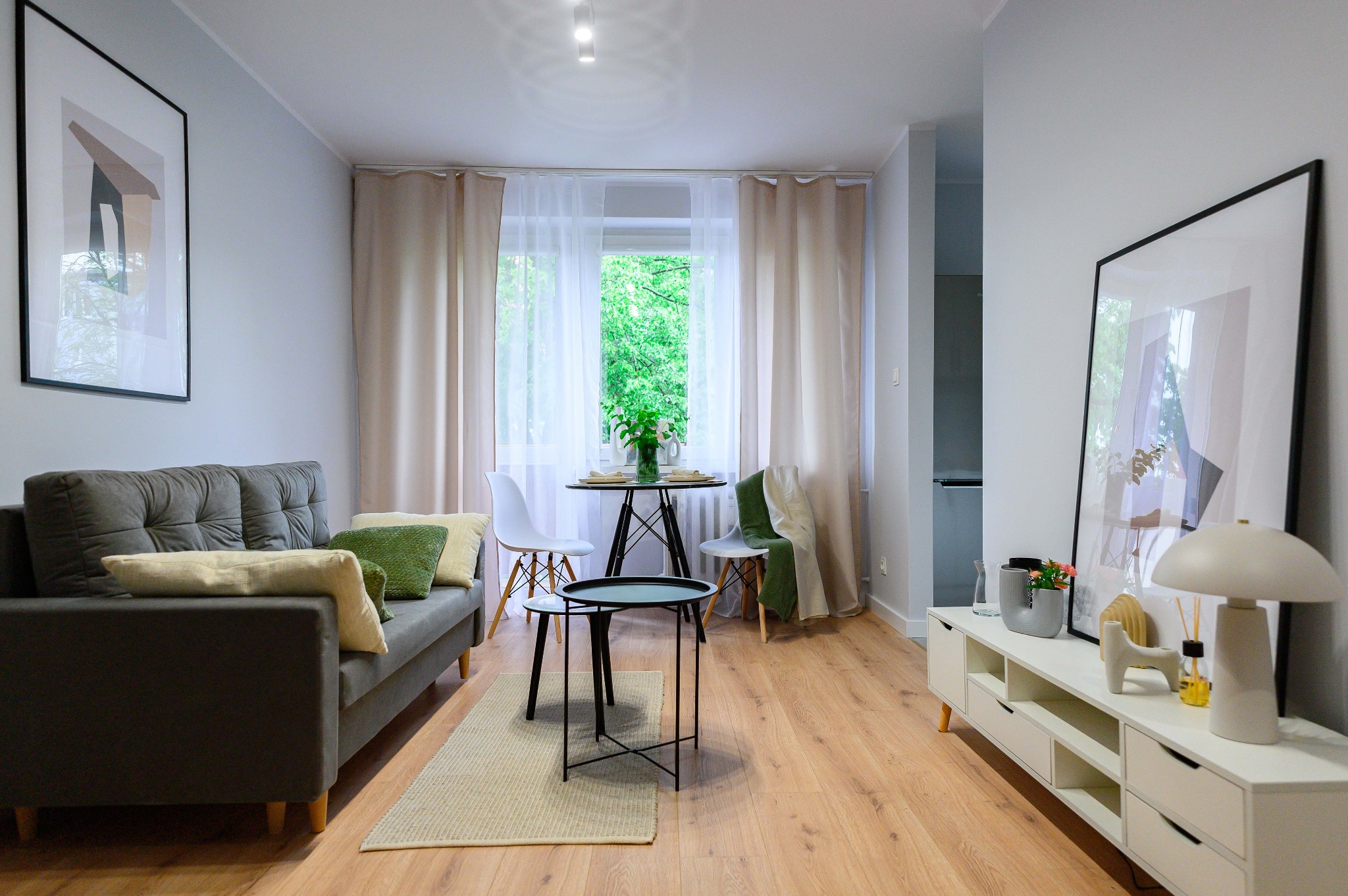 Jasne wnętrze salonu po remoncie: szara sofa z poduszkami, drewniana podłoga, biała komoda z dekoracjami, okno z zasłonami i okrągły stół z krzesłami.