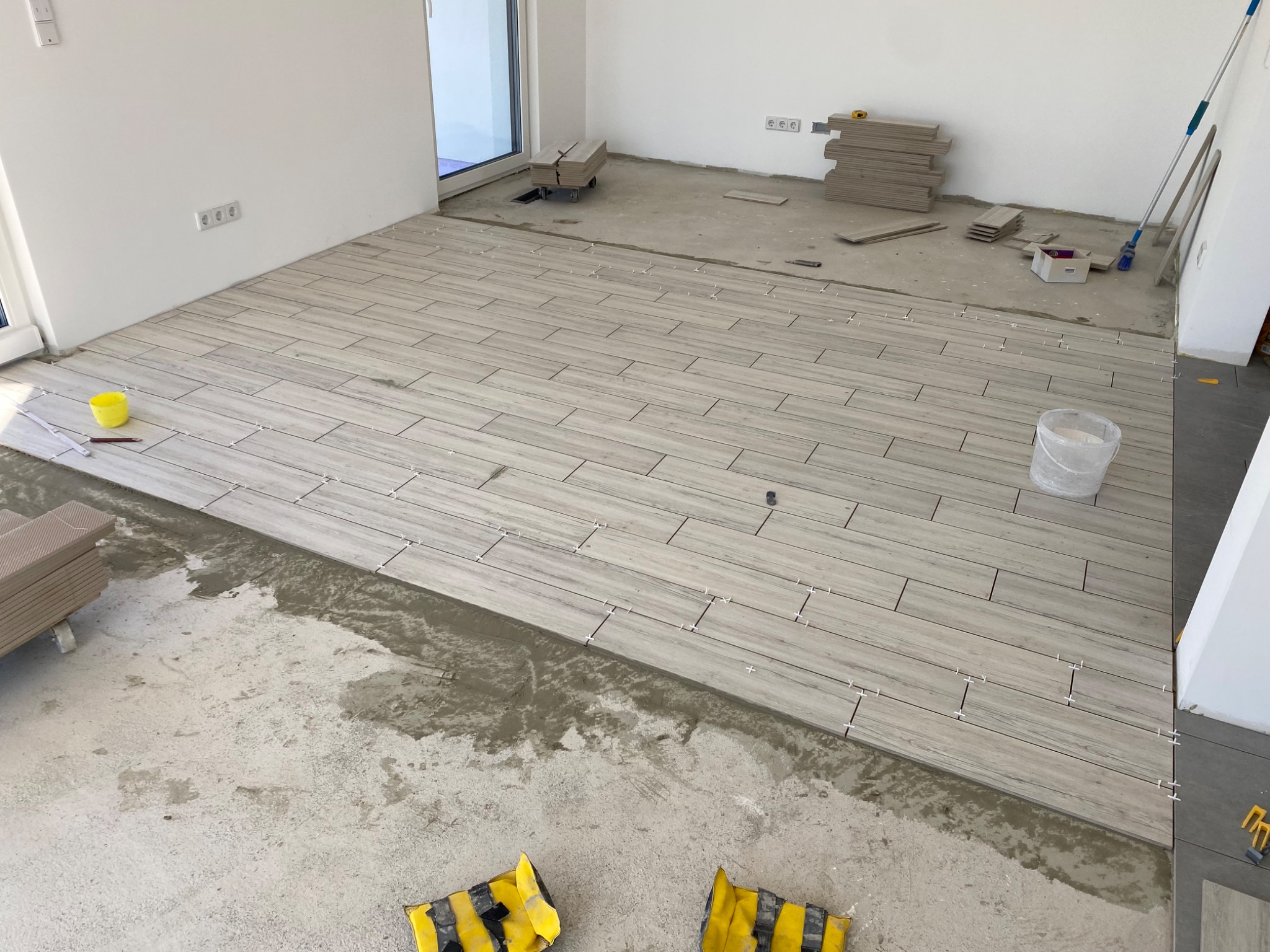 Układanie podłogi z płytek imitujących drewno w jasnym wnętrzu; widoczne płytki, zaprawa, poziomica, wiadro i żółte ochraniacze na kolana.