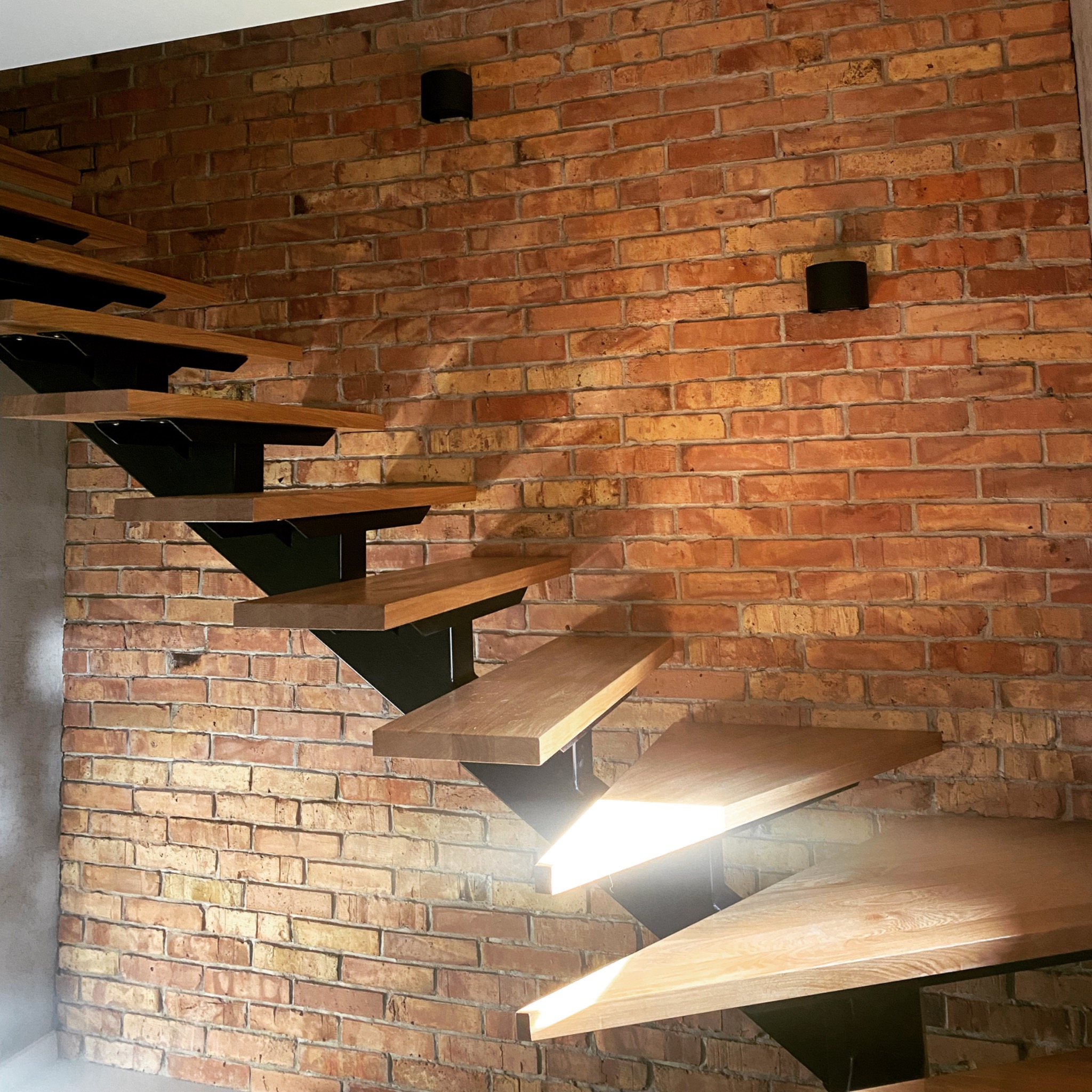 Nowoczesne schody drewniane z czarną metalową konstrukcją na tle ściany z czerwonej cegły i czarnych lamp ściennych.