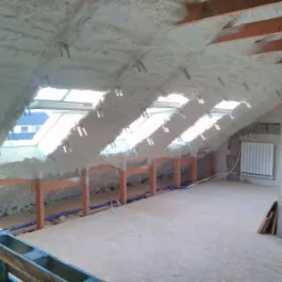 Poddasze z oknami dachowymi w trakcie izolacji pianką, widoczne krokwie i instalacje wodne, surowy stan wykończenia ścian.