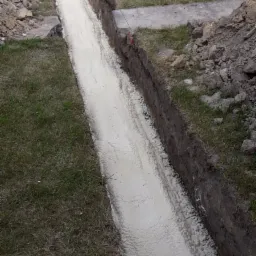 Wąski wykop fundamentowy wypełniony jasną pianą izolacyjną, widoczne ślady trawy i ziemi, tło z ceglanym budynkiem i zielenią.