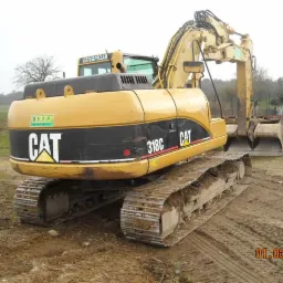 Żółta koparka gąsienicowa Caterpillar 318C stoi na ubitej, błotnistej ziemi. Widoczny profil maszyny z oznaczeniami producenta oraz fragment terenu z cegłami w tle.