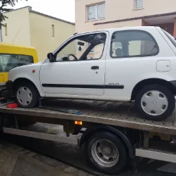 Biały samochód Nissan Micra 16V na platformie żółtej lawety, widok z boku na tle budynków.