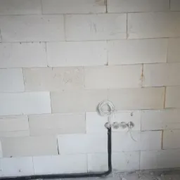 Ściana z betonu komórkowego z trzema wywierconymi otworami, czarnym kablem w peszlu biegnącym w dół i zwiniętym białym przewodem przy otworach.