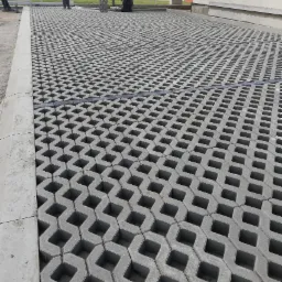 Szara nawierzchnia z ażurowych elementów betonowych, widoczne fragmenty osób układających, obrzeża z kostki.