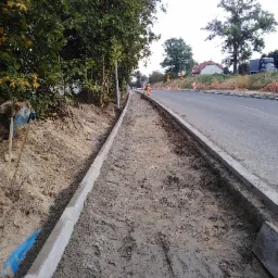 Przygotowanie podłoża pod chodnik z ułożonymi obrzeżami betonowymi wzdłuż drogi, widoczny pracownik w oddali i kopiec ziemi po lewej stronie.