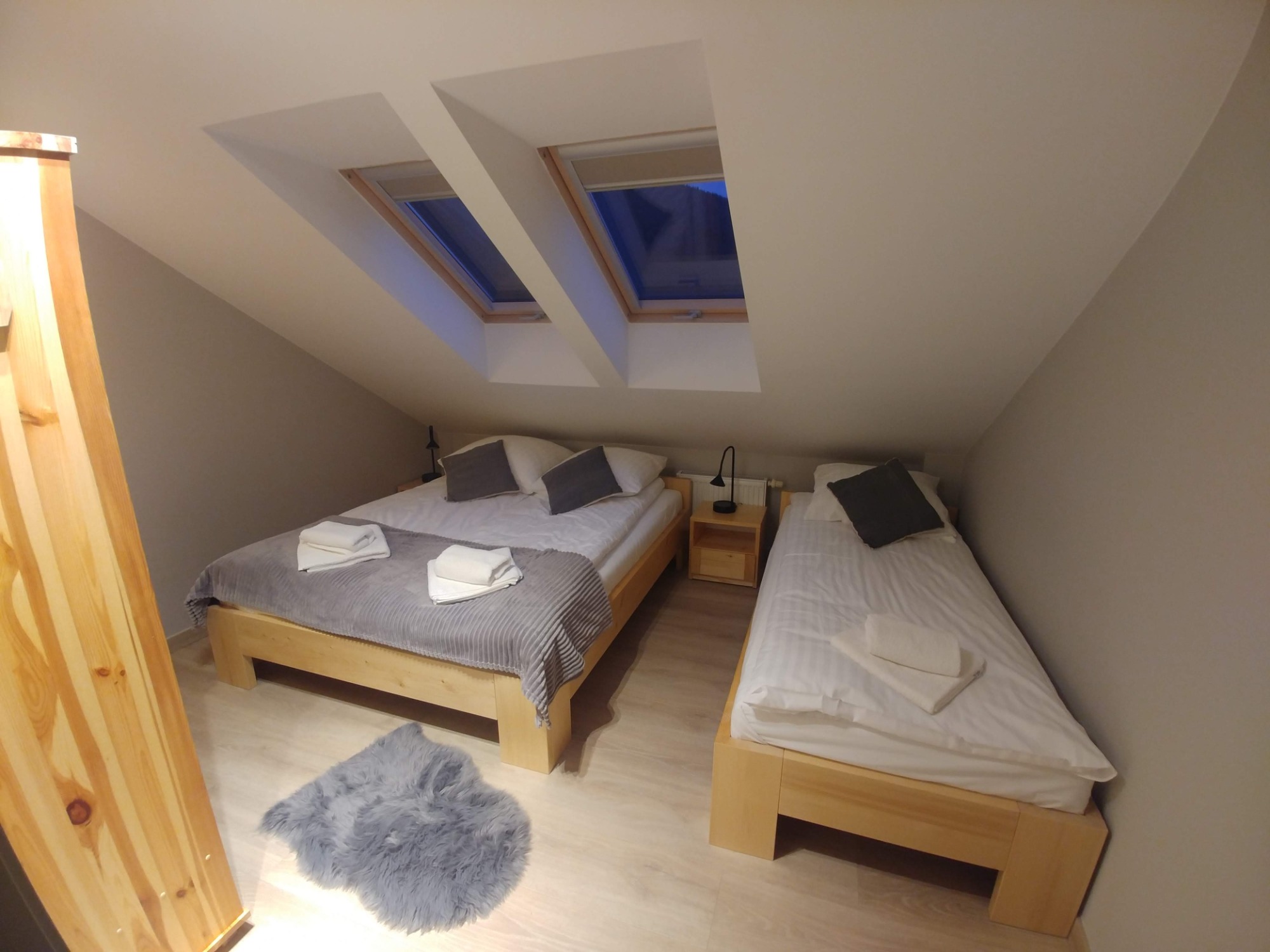 Pokój na poddaszu z dwoma łóżkami z jasnego drewna, szarym dywanikiem i dwoma oknami dachowymi w skośnym suficie.