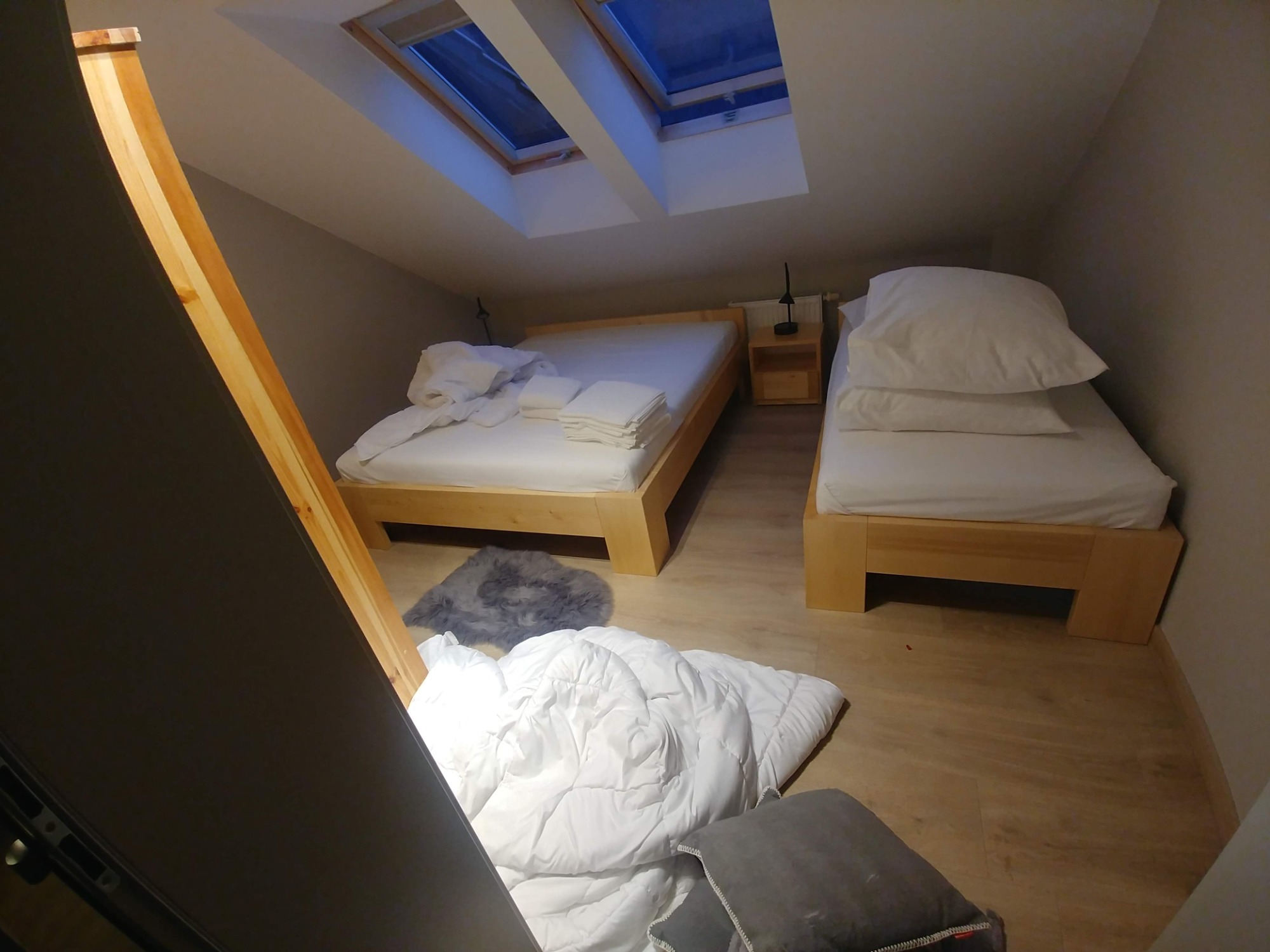 Pokój na poddaszu z dwoma drewnianymi łóżkami, pościelą, dywanikiem i kołdrą na podłodze, oświetlony przez okna dachowe.