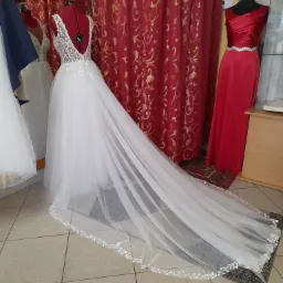 Biała suknia ślubna z koronkowym wykończeniem na czerwonym manekinie krawieckim, długi tren na podłodze z płytek, czerwone zasłony w tle.