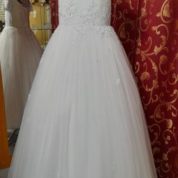 Małgorzata Wal - Biała sukienka komunijna z koronkową górą, prezentowana na manekinie na tle czerwonej zasłony ze złotym wzorem. W lustrze widoczne odbicie sukienki.