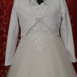 Małgorzata Wal - Elegancka biała sukienka komunijna z tiulowym dołem i bolerkiem zapinanym na ozdobną broszkę, prezentowana na manekinie na tle czerwonej ściany z ornamentami.