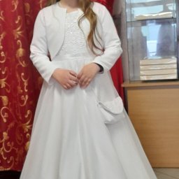 Małgorzata Wal - Dziewczynka w białej sukience komunijnej z bolerkiem i torebką, wianek na głowie, czerwona zasłona w tle.