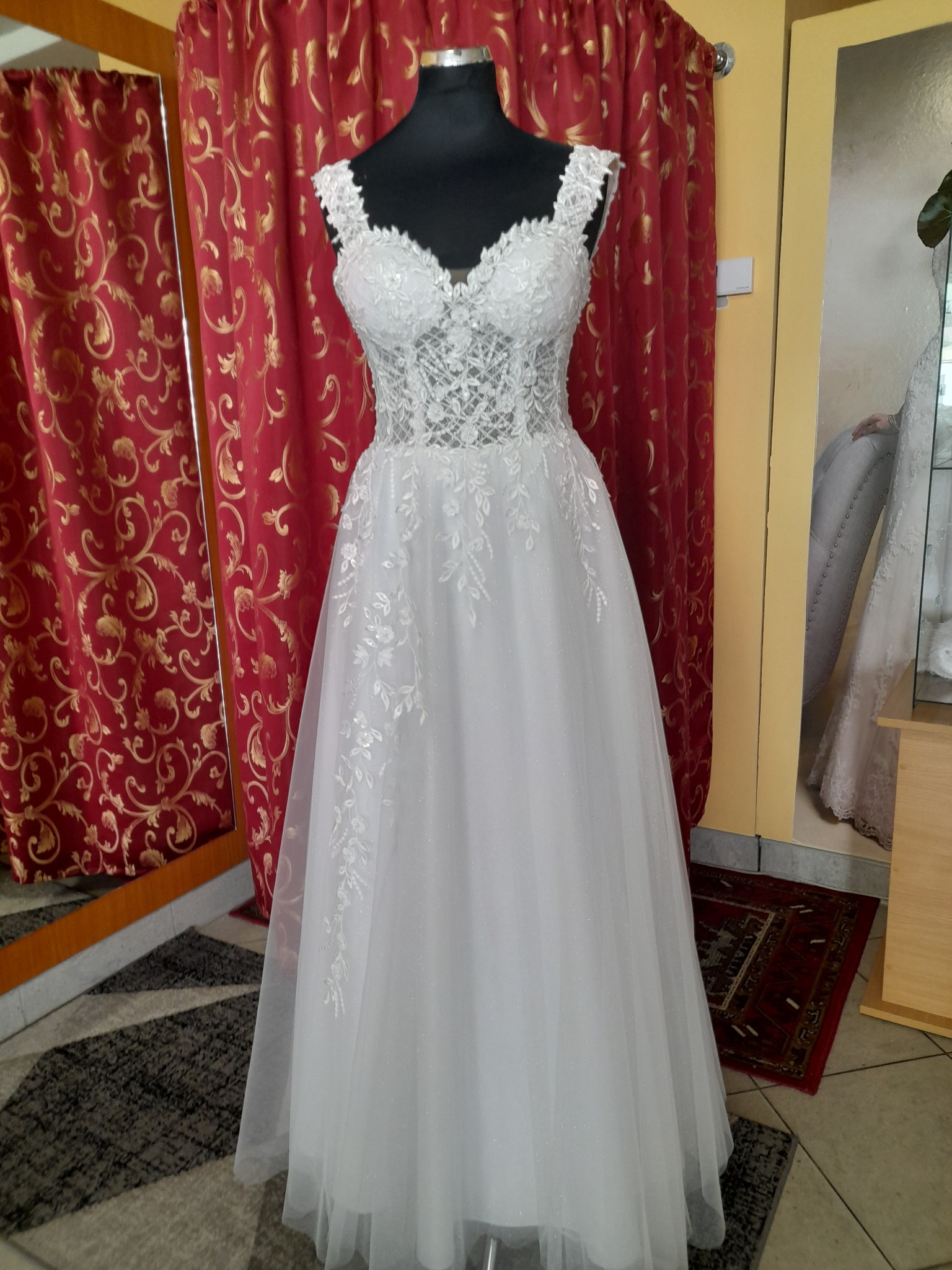 Biała suknia ślubna z koronkową górą i tiulowym dołem, prezentowana na czarnym manekinie w salonie z czerwonymi zasłonami w tle.