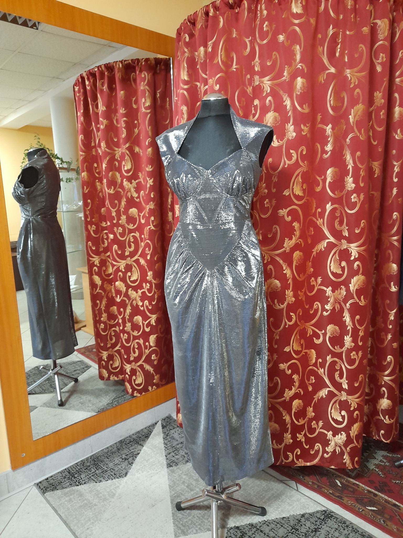 Srebrna, błyszcząca suknia wieczorowa na manekinie krawieckim, odbicie w lustrze, czerwona zasłona w złote wzory w tle.