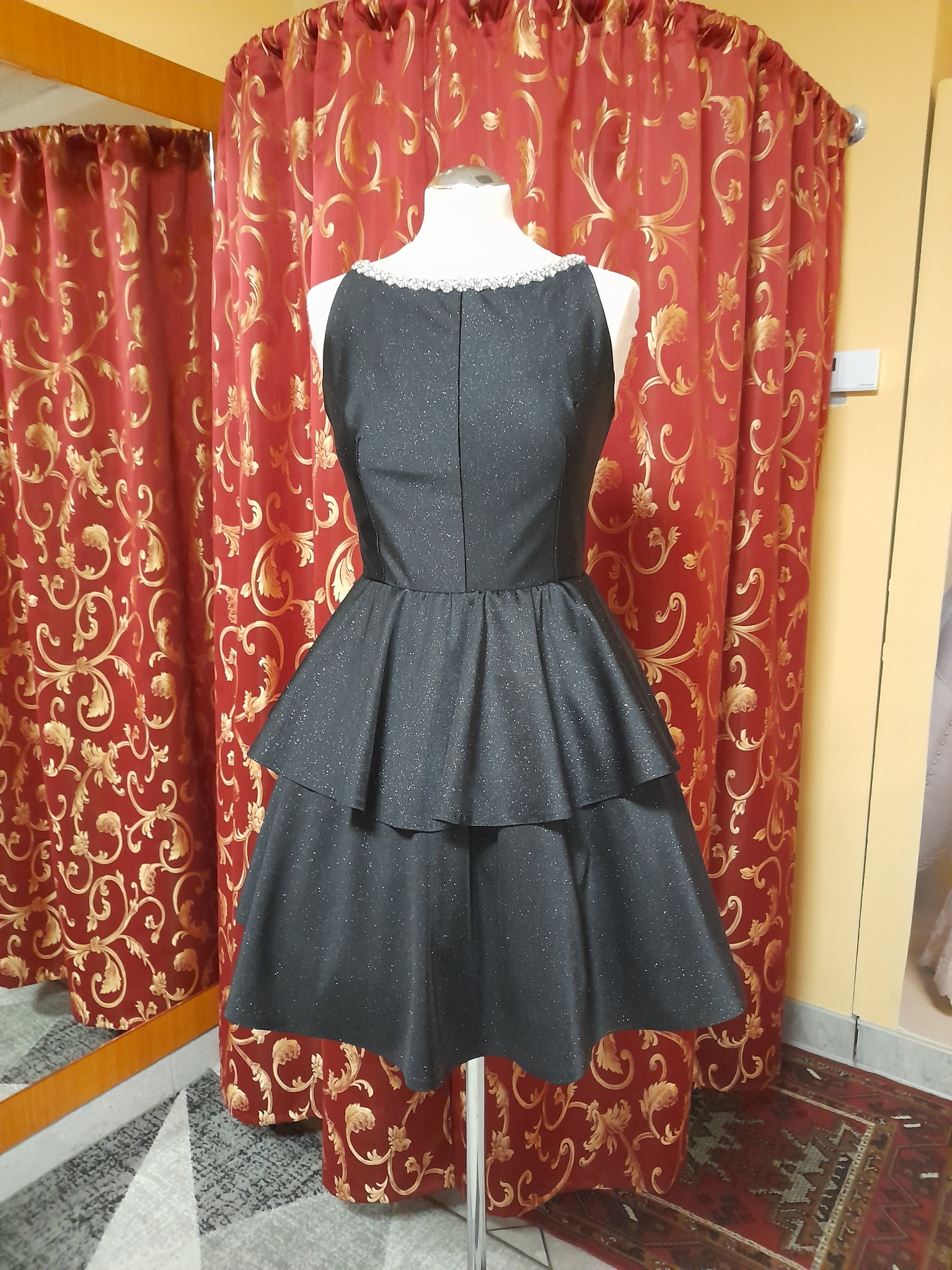 Czarna, błyszcząca sukienka z dwuwarstwową spódnicą na manekinie krawieckim, wykończona koronką przy dekolcie, na tle czerwonych zasłon w złote wzory i lustra.
