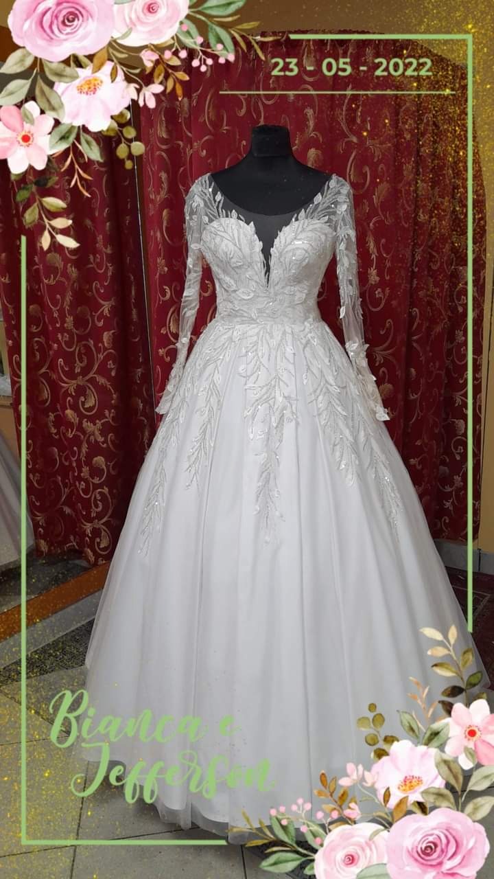 Elegancka suknia ślubna z długimi, koronkowymi rękawami i głębokim dekoltem, prezentowana na manekinie na tle czerwonej, wzorzystej zasłony, z datą 23-05-2022 i imieniem 'Bianca & Jefferson'...