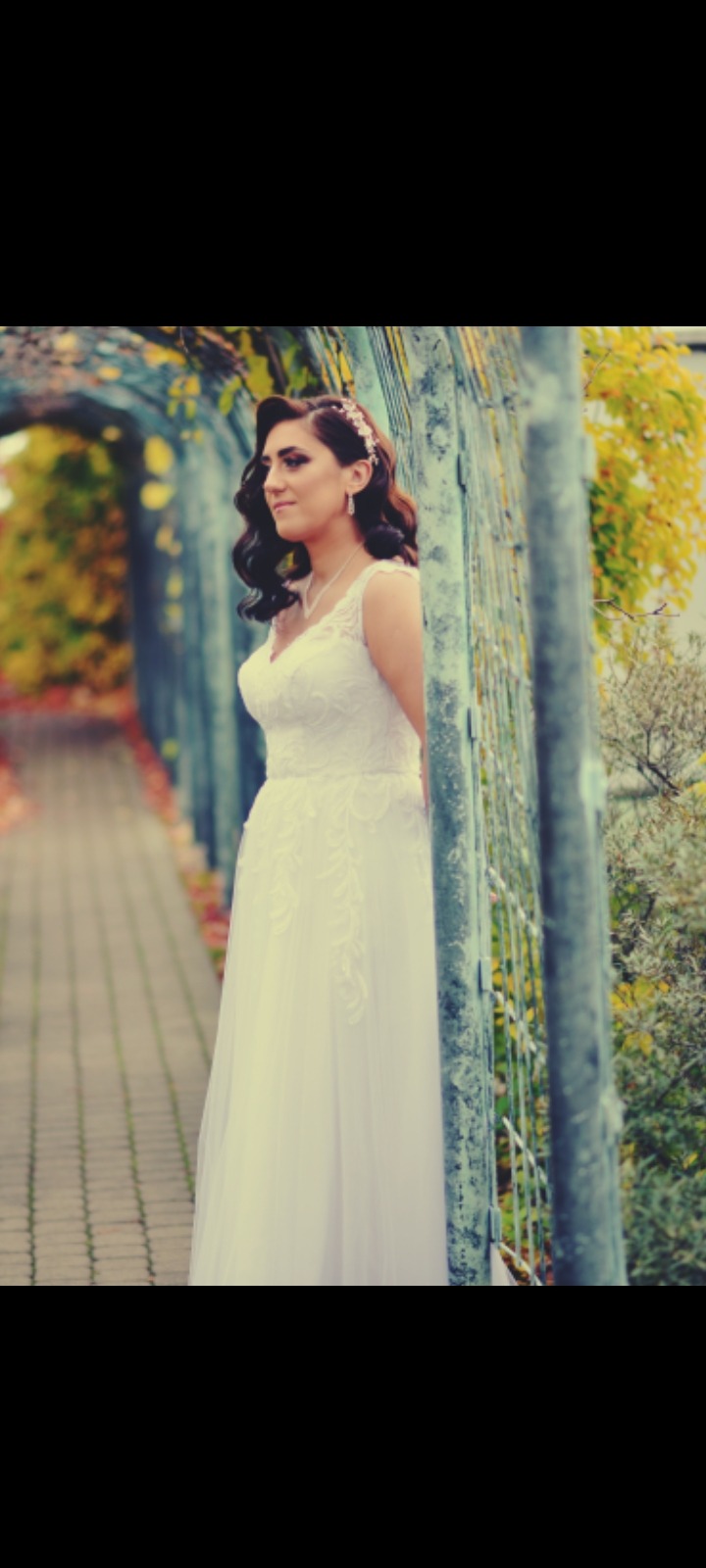 Panna młoda w białej sukni z koronkowymi aplikacjami, pozująca na tle metalowej pergoli z jesienną roślinnością, fryzura z lokami i ozdobą we włosach.