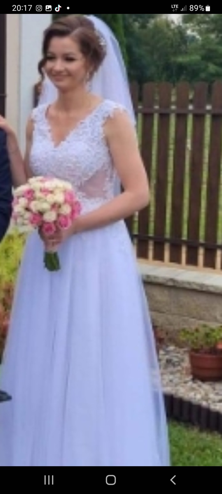 Uśmiechnięta panna młoda w białej sukni ślubnej z koronkową górą i tiulowym dołem, trzyma bukiet różowych i białych róż, widoczny fragment drewnianego płotu i doniczka z kwiatami w tle.