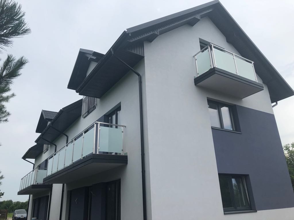 Nowoczesny budynek wielorodzinny z balkonami zabezpieczonymi szkłem i metalem, widok z dołu na szaro-białą elewację w pochmurny dzień.
