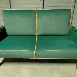 Dwuosobowa sofa w kolorze butelkowej zieleni, obita welurem i wykończona złotą lamówką, stoi na drewnianych nogach na tle szarej ściany.