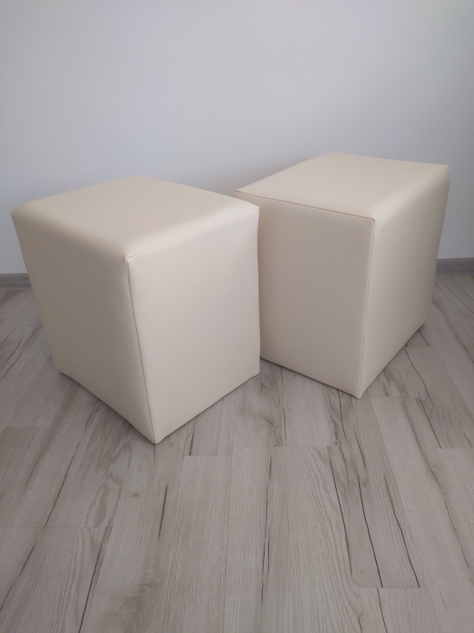 Dwa identyczne, kremowe pufy w kształcie sześcianu, obite gładką tkaniną, stoją na jasnej podłodze imitującej drewno, na tle białej ściany.