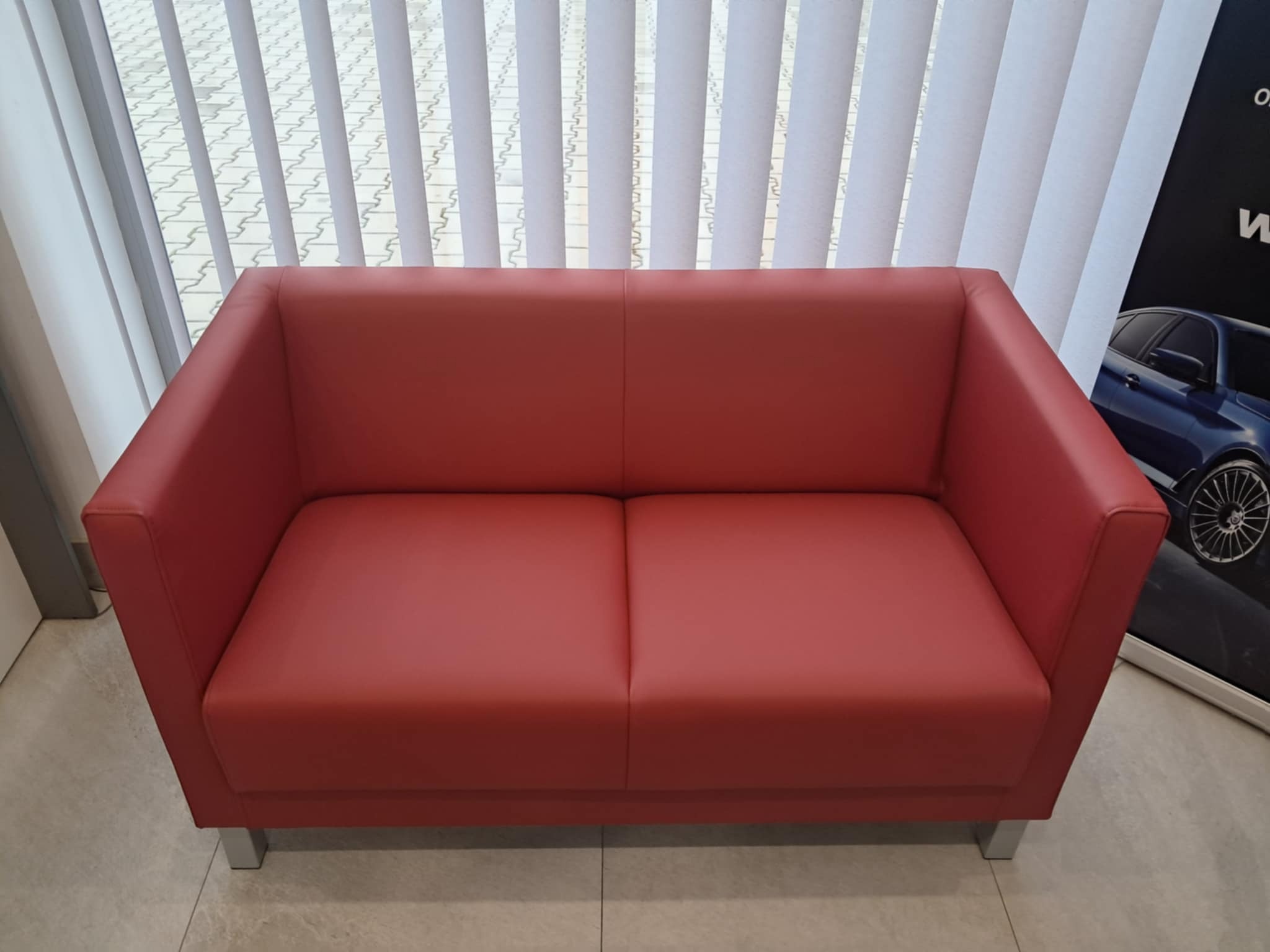 Nowoczesna dwuosobowa sofa w odcieniu koralowej czerwieni, obita gładką skórą ekologiczną, z minimalistycznymi, chromowanymi nóżkami, ustawiona na tle pionowych żaluzji i plakatu z samochodem.