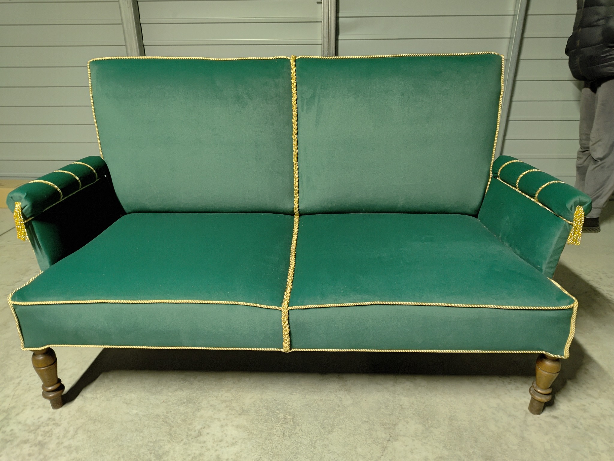 Dwuosobowa sofa w kolorze butelkowej zieleni, obita welurem i wykończona złotą lamówką, stoi na drewnianych nogach na tle szarej ściany.