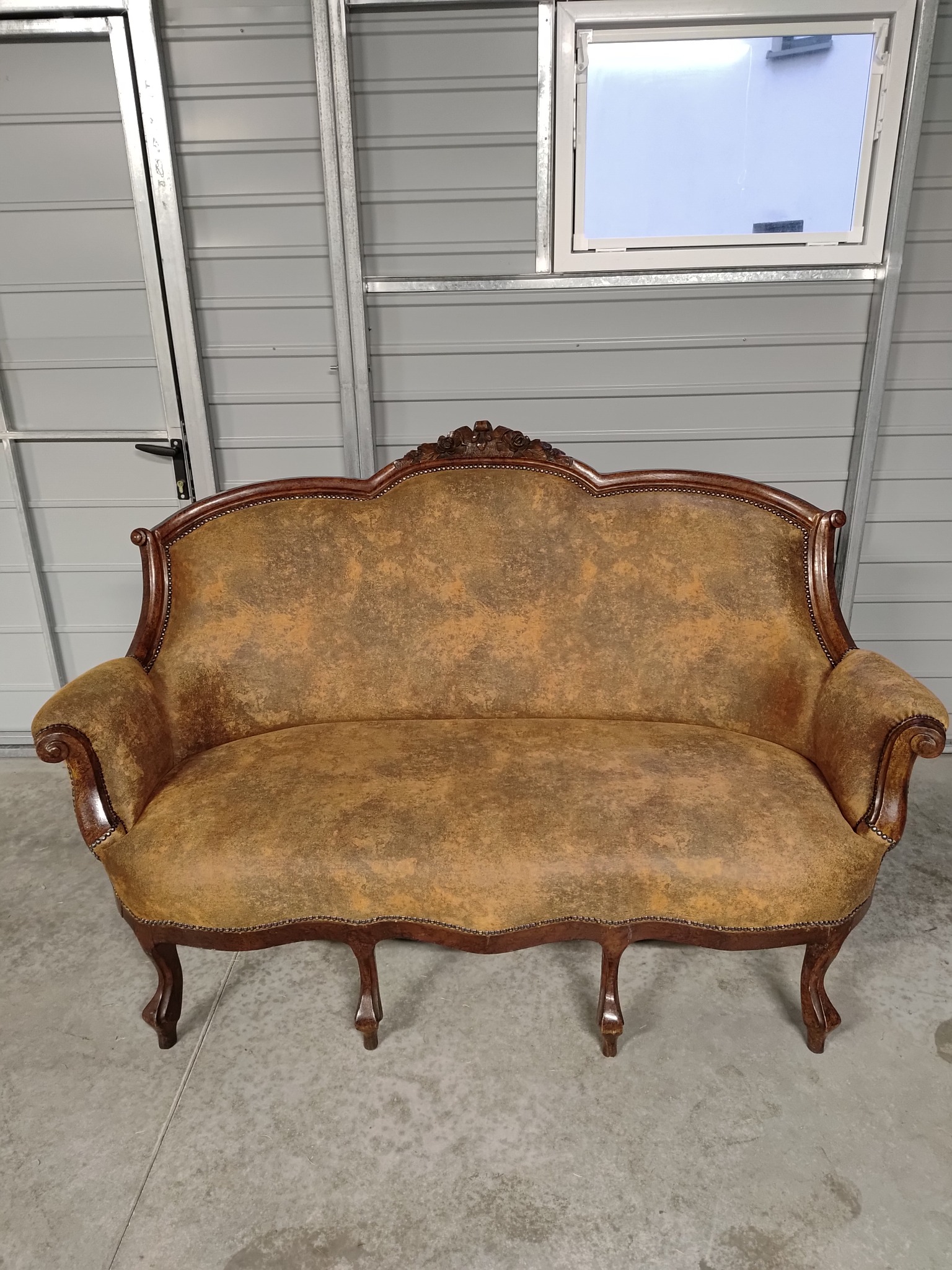 Antyczna sofa z brązową, wzorzystą tapicerką, drewnianą ramą zdobioną rzeźbieniami i gwoździami tapicerskimi, sfotografowana w pomieszczeniu z blachy falistej z oknem.