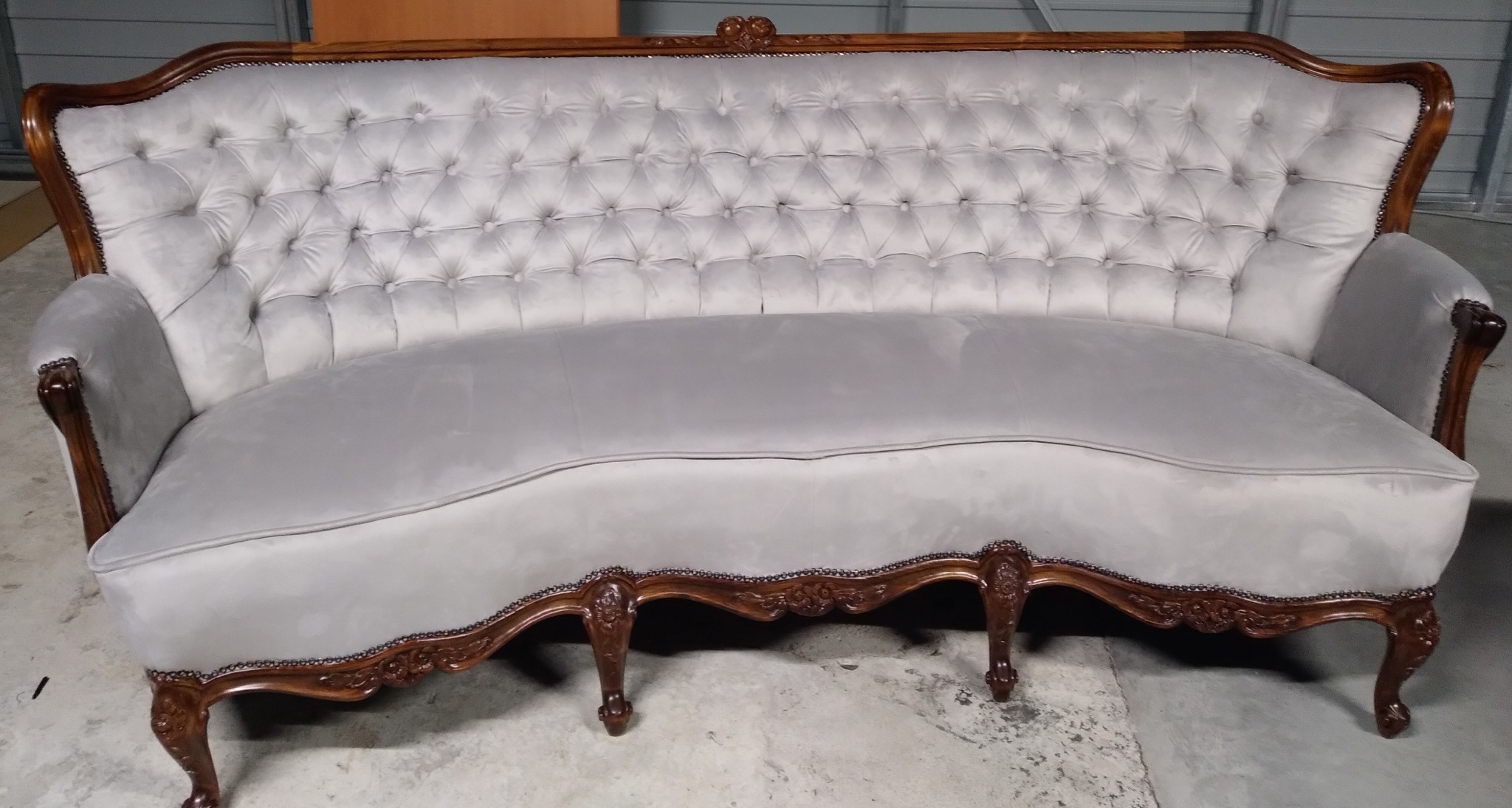 Elegancka, jasnoszara sofa w stylu Ludwika Filipa z drewnianą ramą o bogatych zdobieniach i pikowanym oparciem, ustawiona na betonowej podłodze.