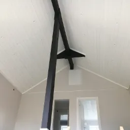 Wnętrze domu z białym drewnianym sufitem, wpuszczonymi lampami, szarobeżowymi ścianami i czarną, drewnianą konstrukcją nośną.