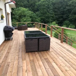 Drewniany taras z jasnych desek z meblami ogrodowymi z rattanu i grillem obok białego budynku, widok na zielony ogród i las.