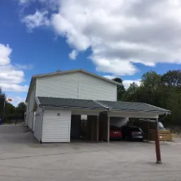 Biały budynek z zadaszonym parkingiem na kilka samochodów, widoczne trzy zaparkowane auta, w tle budynek z wieżą i flaga na maszcie, błękitne niebo z chmurami.