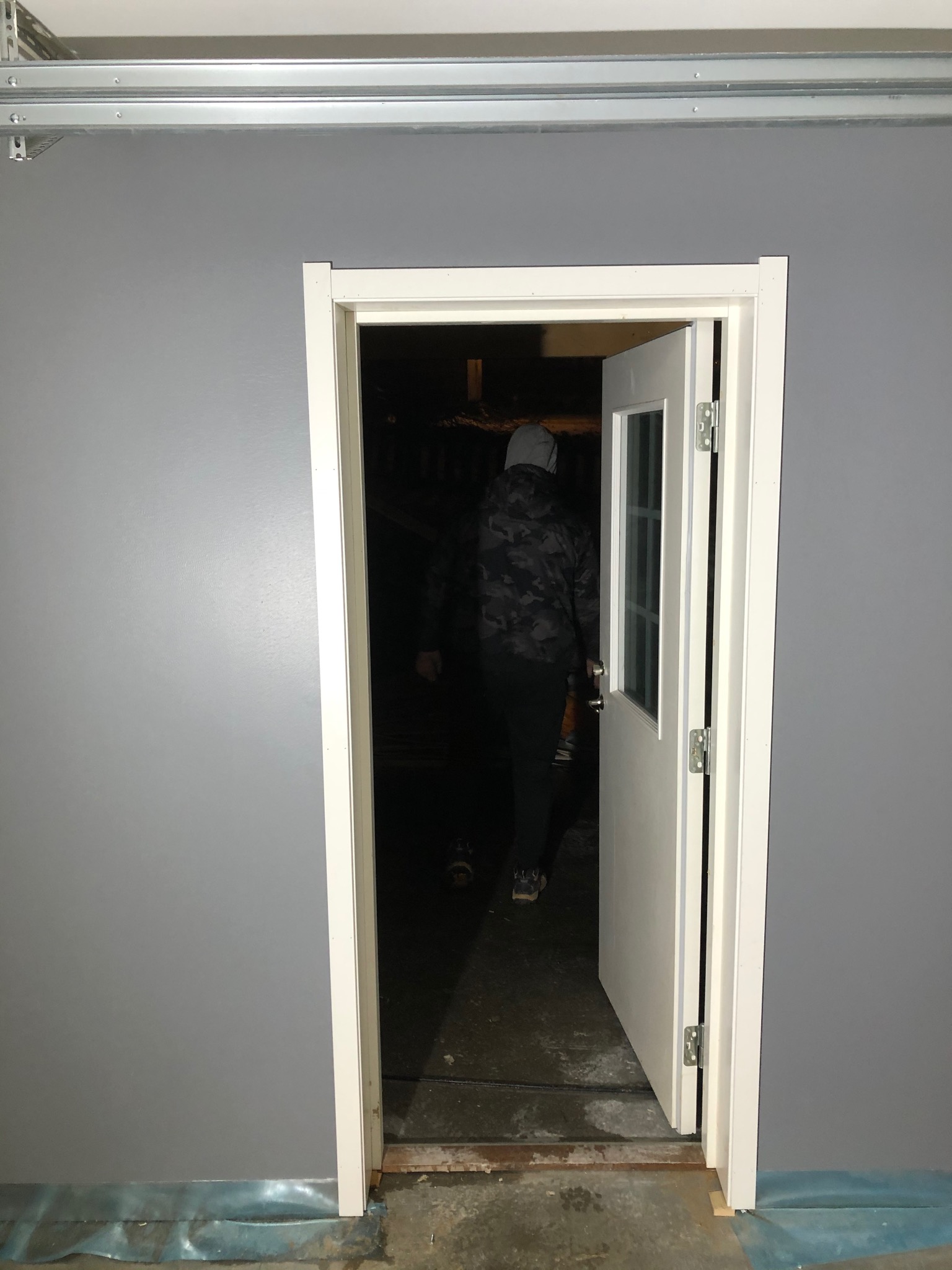 Szara ściana z nowo osadzonymi białymi drzwiami z szybą; osoba w kurtce moro wchodzi do pomieszczenia.