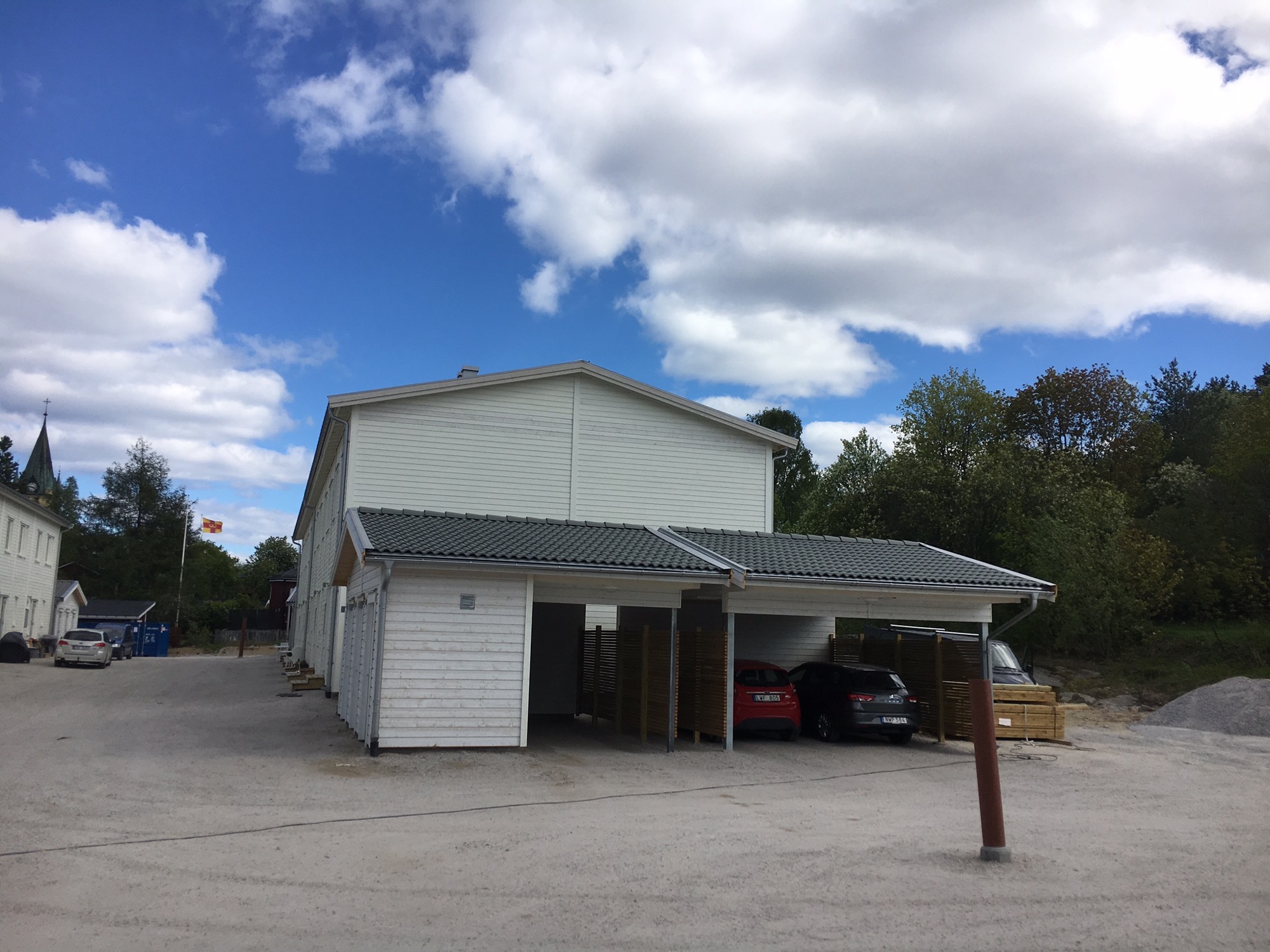 Biały budynek z zadaszonym parkingiem na kilka samochodów, widoczne trzy zaparkowane auta, w tle budynek z wieżą i flaga na maszcie, błękitne niebo z chmurami.