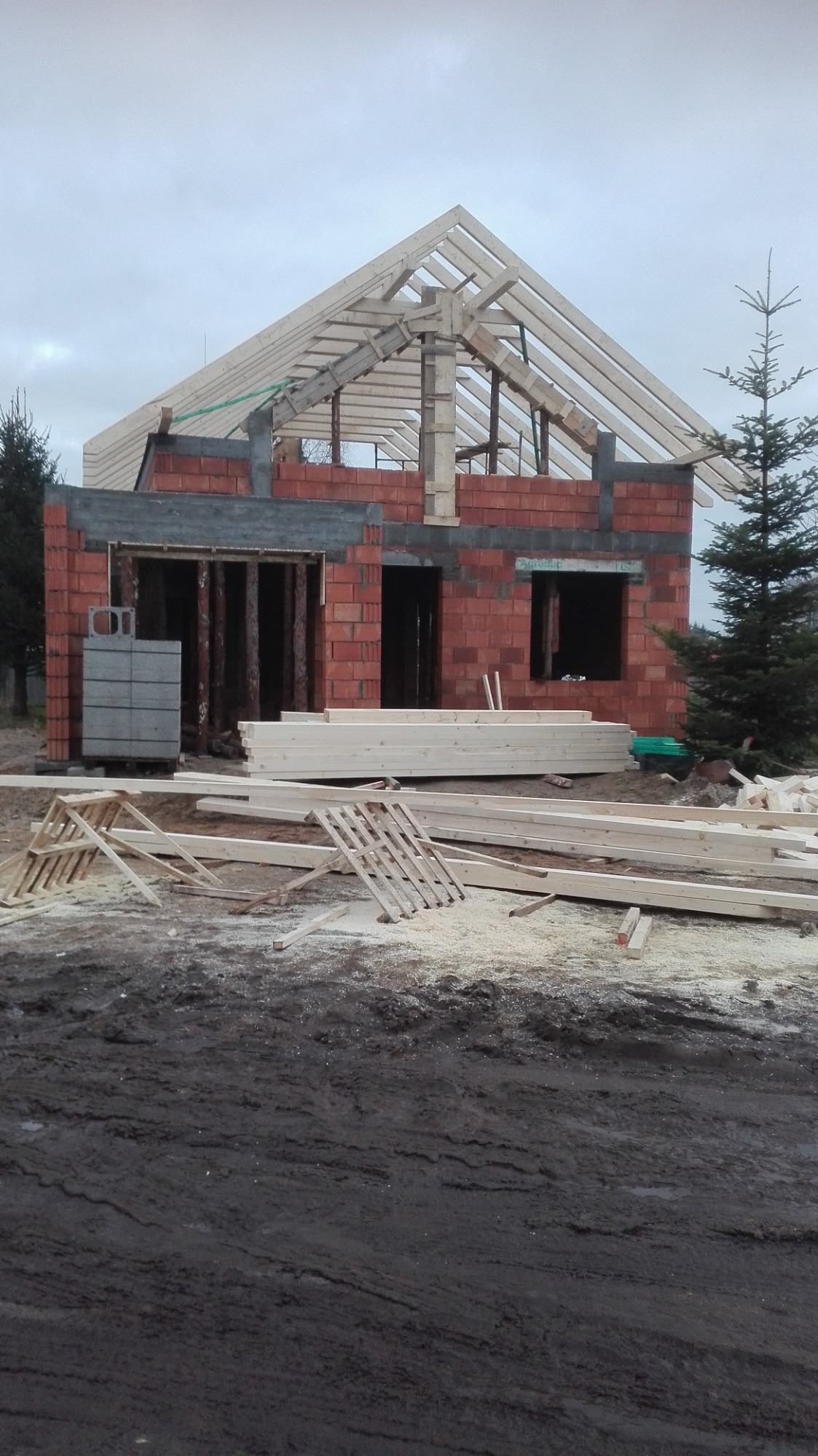 Budowa domu w stanie surowym otwartym: widoczne mury z czerwonej cegły, drewniana konstrukcja dachu, stosy tarcicy budowlanej i bloczki betonowe na błotnistym terenie.