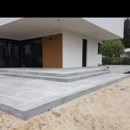 Nowoczesny dom z szarą kostką brukową tworzącą taras i schody wejściowe, widoczne ziarna piasku przy krawędziach kostki.