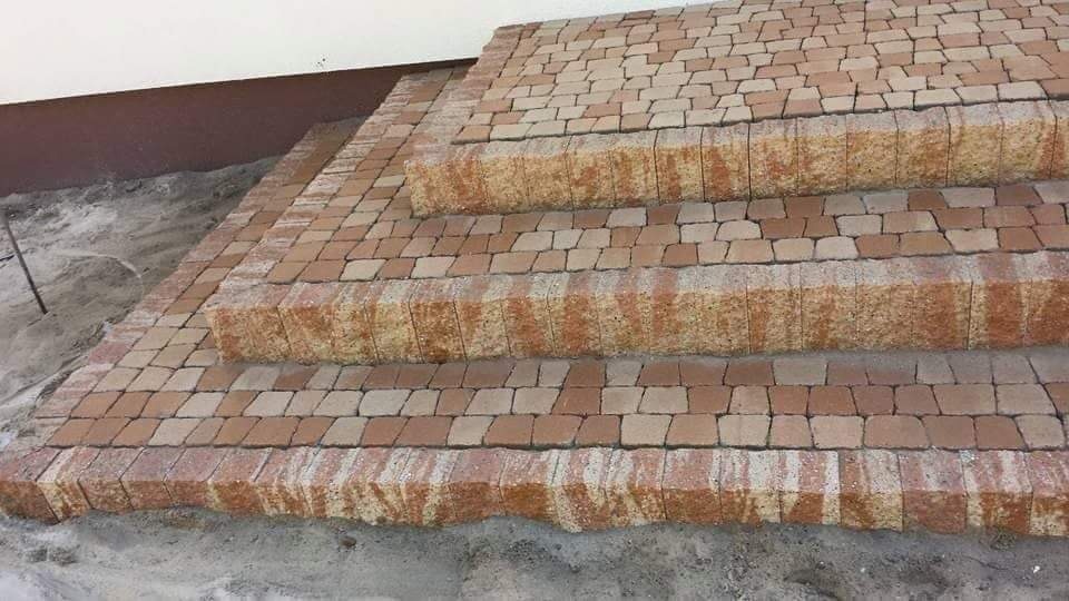 Schody wejściowe wyłożone czerwonobrązową kostką brukową na podsypce piaskowej, widoczne niedociągnięcia przy ścianie budynku.