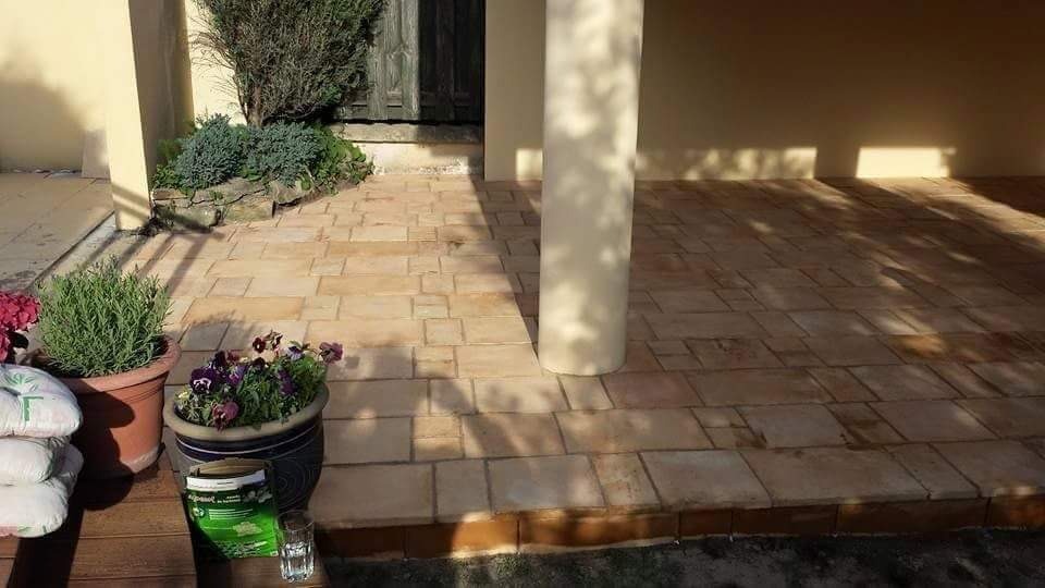 Wyłożony kostką brukową taras z kolumną, doniczkami z kwiatami i ziołami, oraz workami z materiałem budowlanym, w słoneczny dzień.