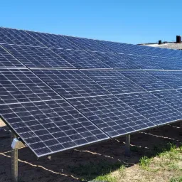 Instalacja paneli fotowoltaicznych na ziemi, widoczne metalowe konstrukcje wsporcze, w tle dom z ciemnym dachem i metalowe ogrodzenie.