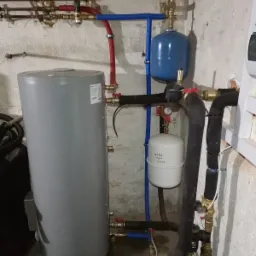 Szary zbiornik akumulacyjny ciepłej wody użytkowej z podłączonymi rurami miedzianymi, stalowymi i z tworzywa sztucznego w kotłowni z surowymi ścianami.