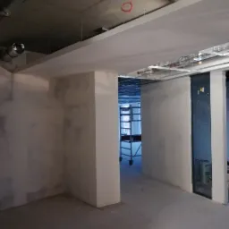 Surowe wnętrze w Krakowie w trakcie remontu, widoczne ściany po gładziowaniu, sufit z instalacjami i wisząca rękawica robocza, w tle rusztowanie.