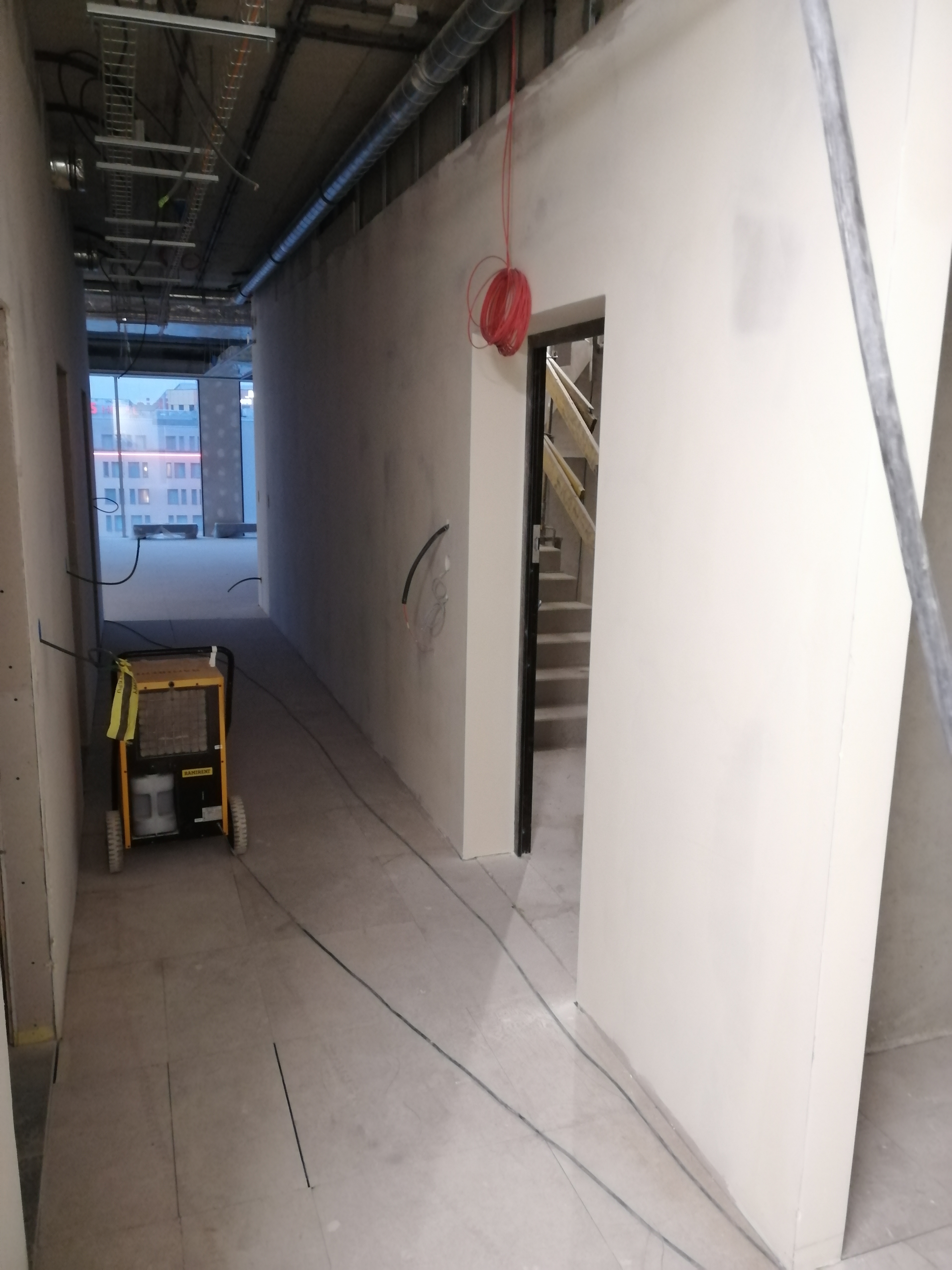 Korytarz w trakcie prac wykończeniowych, ściany pokryte gładzią, widoczny agregat grzewczy Ramirent, zwisający czerwony kabel oraz fragment schodów w głębi
