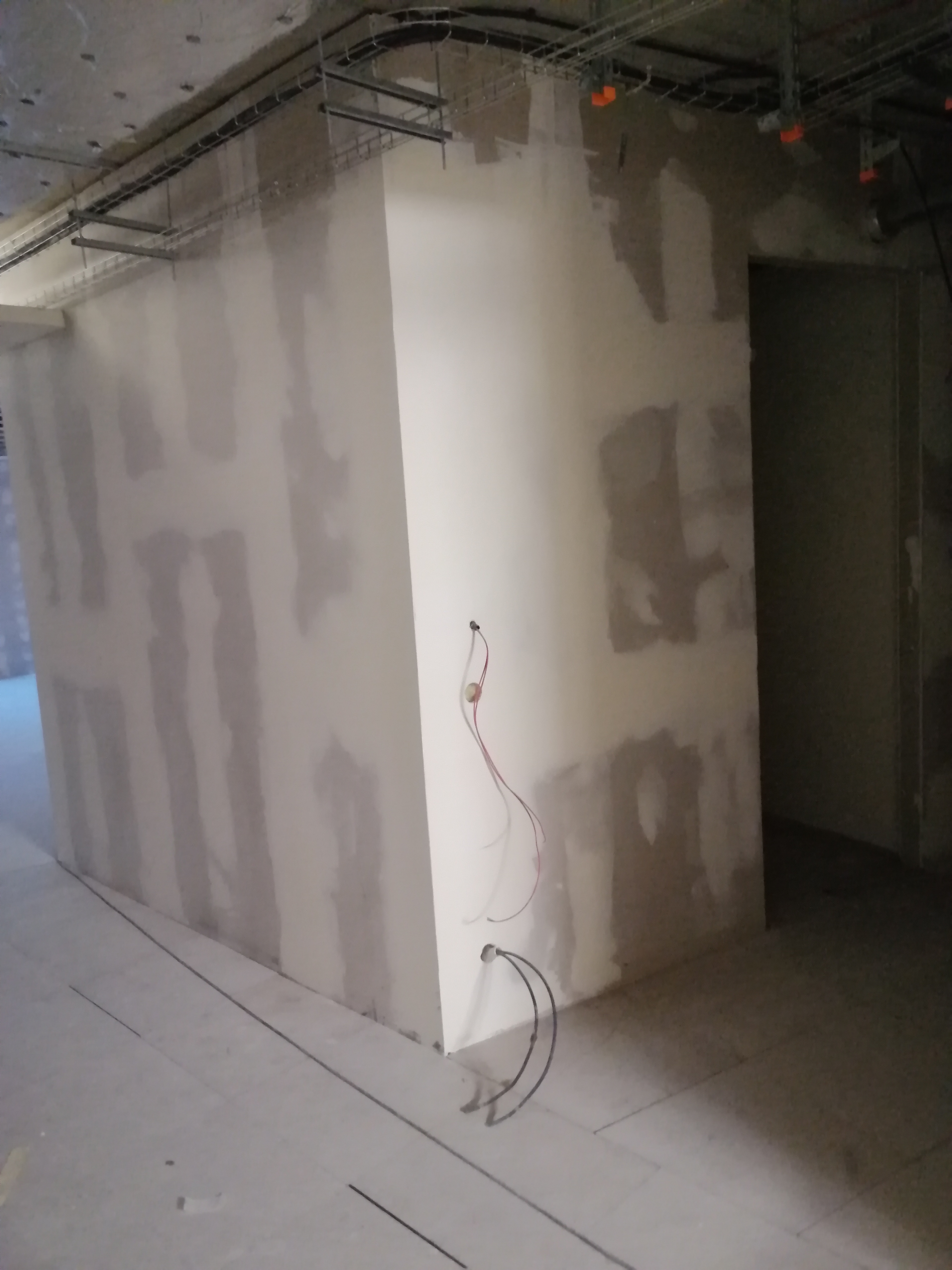 Wnętrze w trakcie prac wykończeniowych: ściany z widocznymi śladami po łatach gipsowych, częściowo wygładzone, wystające przewody elektryczne, widoczna konstrukcja pod sufitem.