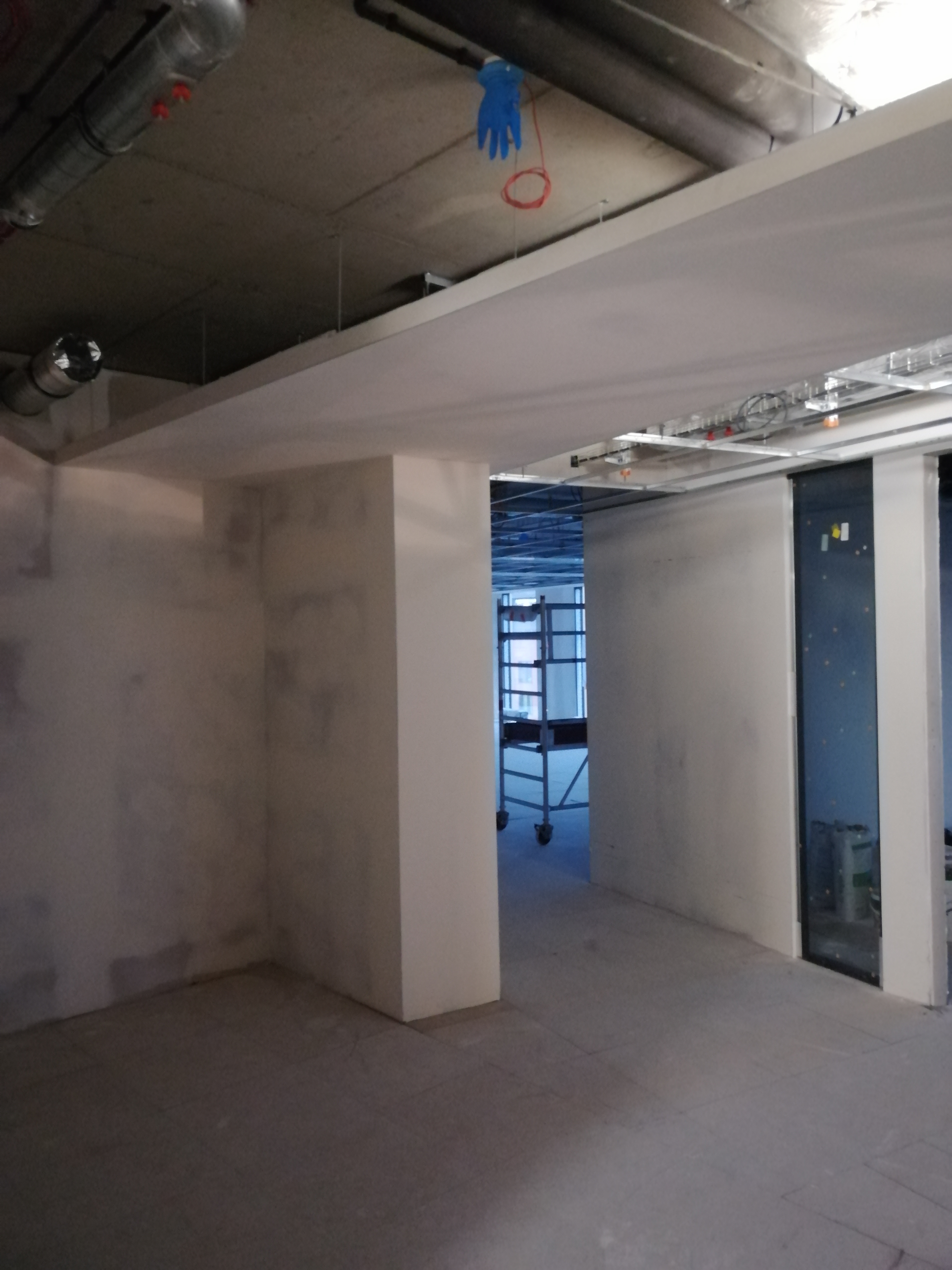 Surowe wnętrze w Krakowie w trakcie remontu, widoczne ściany po gładziowaniu, sufit z instalacjami i wisząca rękawica robocza, w tle rusztowanie.